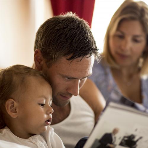 Man och kvinna med litet barn läser bok i hotellsäng.